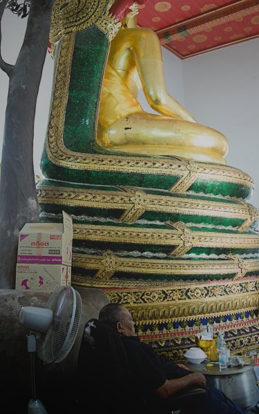 L'envers du décor. Derrière le grand Bouddha assis, un garde somnole sur une chaise. Devant lui, un tambour en métal sert de petite table ou se trouve de la nourriture et des boissons, derrière lui un ventilateur et ce qui semble être une grosse boite de désodorisant .