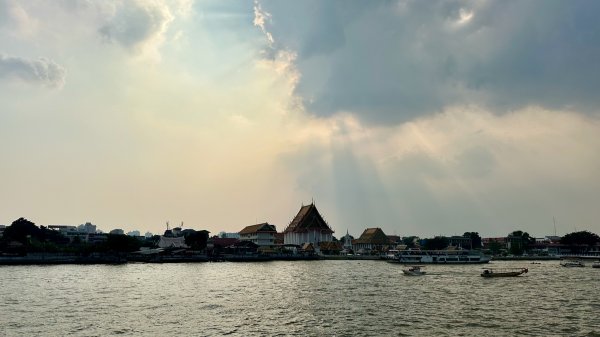 Une vue sur les berges du fleuve avec des nuages à travers desquels filtre des rayons de soleil. Un temple et des bâtiments traditionnels sont visibles sur l'autre rive.