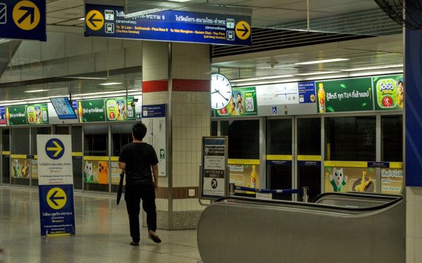 Photographie d'une station de métro à Bangkok, panneaux bleu vif, flèches de directions noires sur fond jaune, publicités et éclairage crus. Un homme de dos marche avec un parapluie à la main.