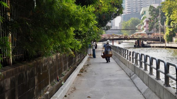 Une personne de dos marchant sur le quai et portant avec un balancier deux grosses gamelles sur son épaules.