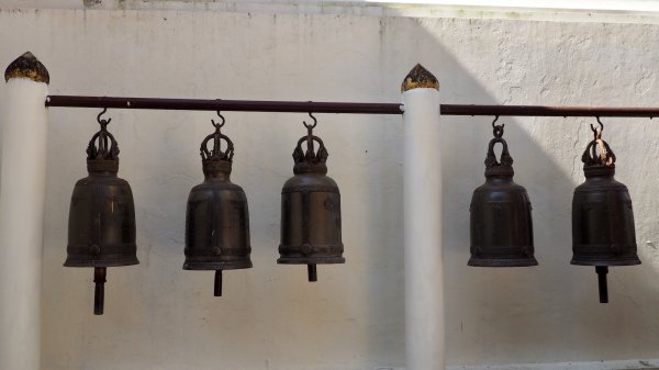 Cinq cloches anciennes suspendues à une barre en bois devant un mur blanc.