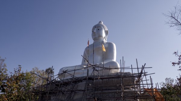 Un bouddha tout blanc en haut de la montagne. Des échafaudages en bambou sont dressés devant la base et deux petits drapeaux flottent devant lui.