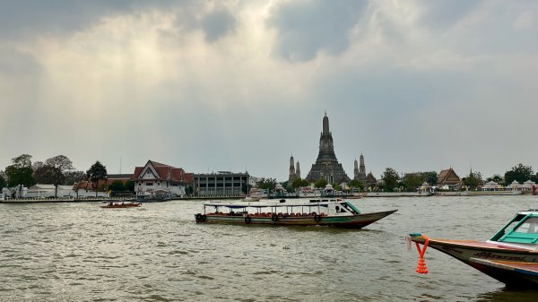 Vue sur le temple Wat Arun depuis les berges du fleuve. Ciel nuageux et bateaux touristiques colorés qui traversent.