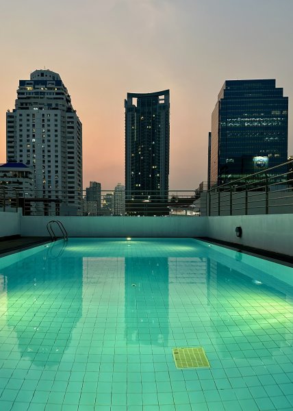 Vue de la piscine sur le toit en fin de journée. Elle fait face à trois grandes tours illuminées. L'eau de couleur vert bleu est éclairée par des spots immergés.
